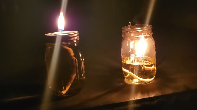 A mason jar closed oil lamp or candle next to a mason jar open oil lamp in a dark room.