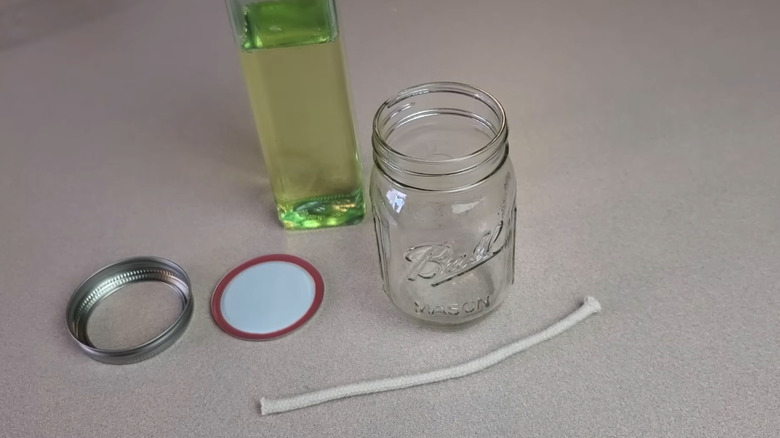 A mason jar, mason jar lid and ring, cotton wick, and a bottle of oil on a white table.
