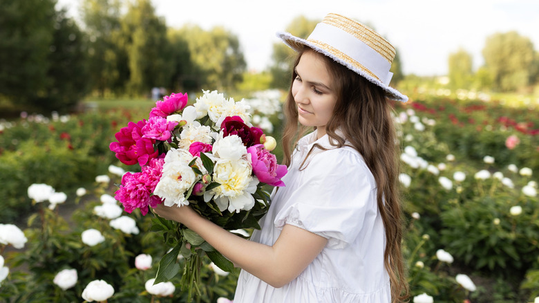 A young person is standing in a garden full of peonies, holding a mixed bouquet of different colors of the flower