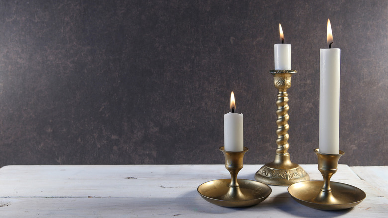 Three brass candlesticks with burning candles are arranged on a white wooden table.