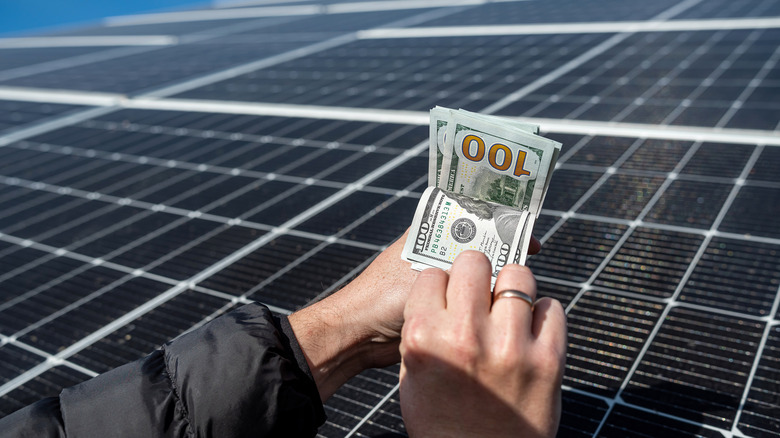 Hands holding multiple $100 bills in front of solar panels
