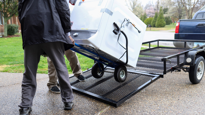 Two people using a dolly to move a washing machine off of a trailer