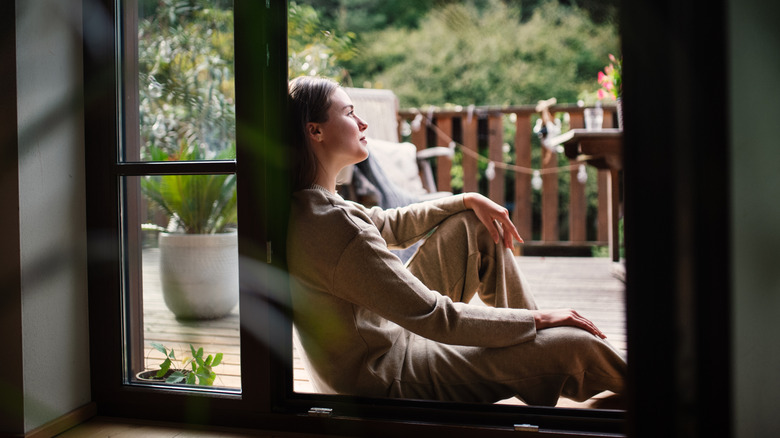 A person rests near on their balcony, resting on the window frame, with potted plants and greenery in the background