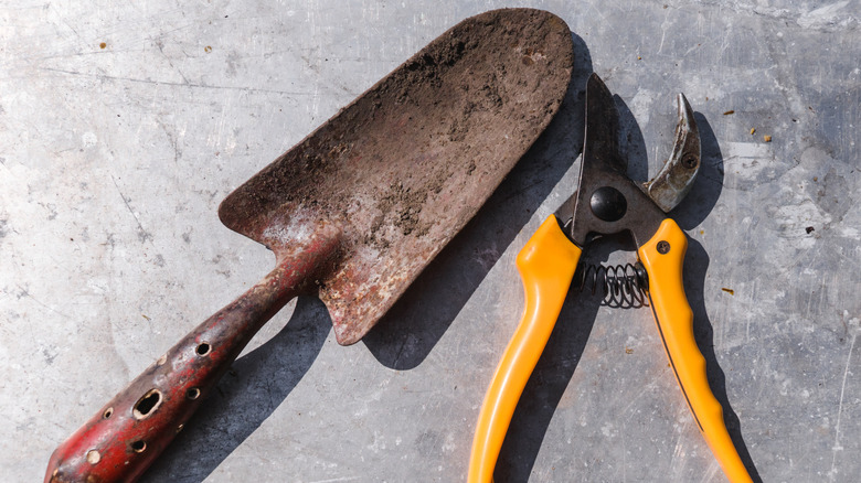 Dirty red garden shovel and yellow pruner on gray floor.