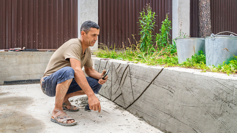 A man crouches beside an old, crumbling, cracking concrete retaining wall in a backyard.