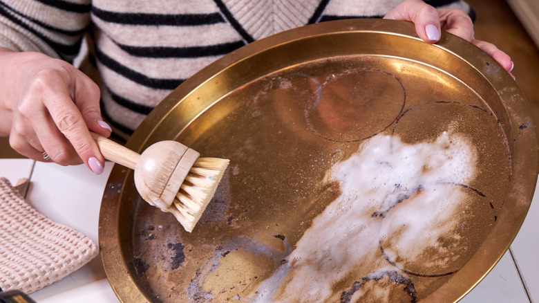 A person cleaning a copper tray with white paste and a wooden brush