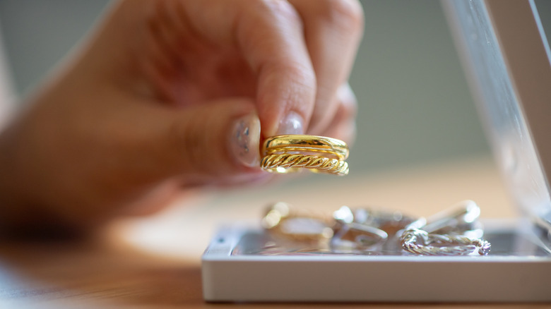 A hand holds a gold ring above other gold and silver rings in a jewelry box.