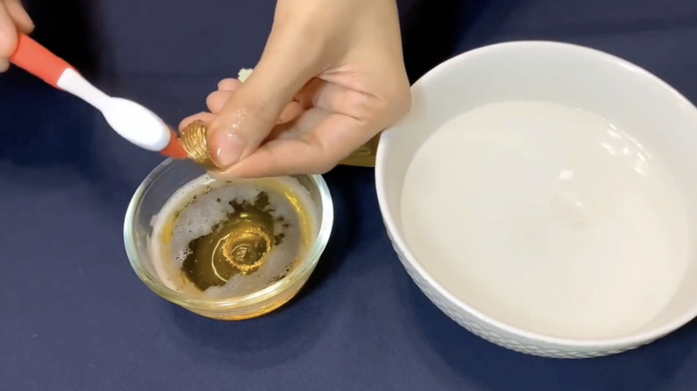 A hand scrubs a gold ring with a toothbrush while holding it above a small glass bowl of beer.