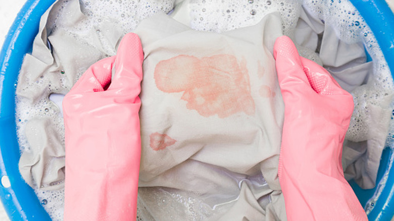 A person with pink gloves washing clothing with a red stain in a bucket