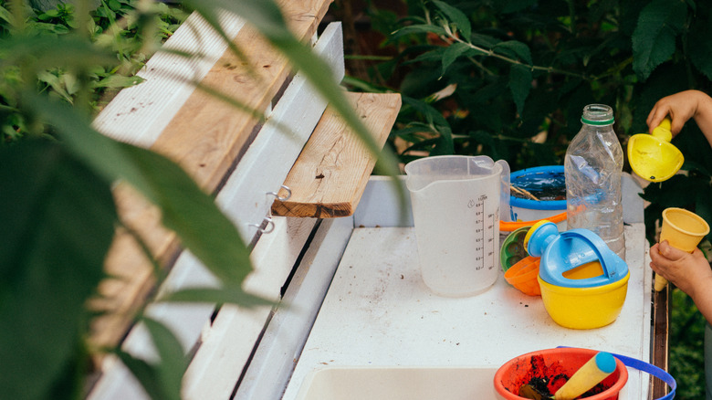 A child with yellow and blue plastic toys playing in a white and wood colored mud kitchen.