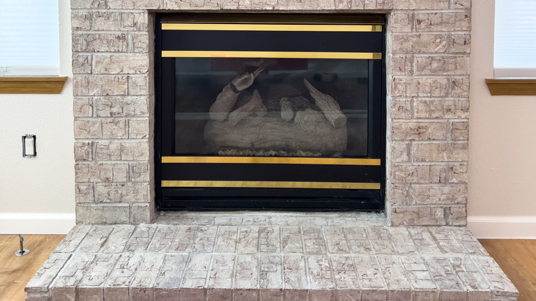 Lightly whitewashed brick fireplace with a raised hearth and a black firebox featuring gold horizontal trim, centered between two windows with white shades.