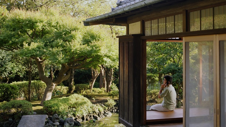A person talking on a cellphone while sitting on the porch of a modern Japanese home in front of a lush garden area