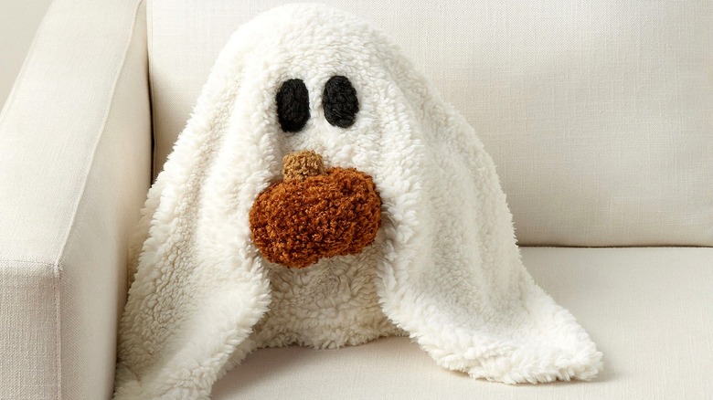 Pottery Barn Pillow in the shape of a ghost holding a pumpkin, sitting on a beige couch.