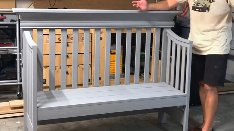 A man in a tan shirt standing next to a crib that has been turned into a bench.