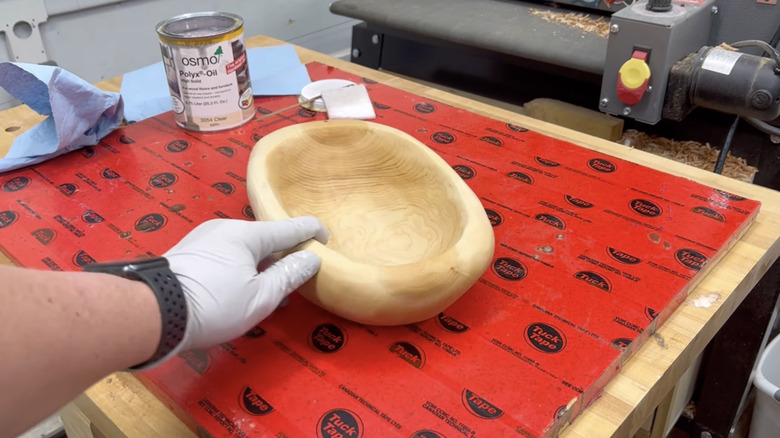 Person's hand wearing a glove and pointing at a finished handmade wood bowl on a work bench