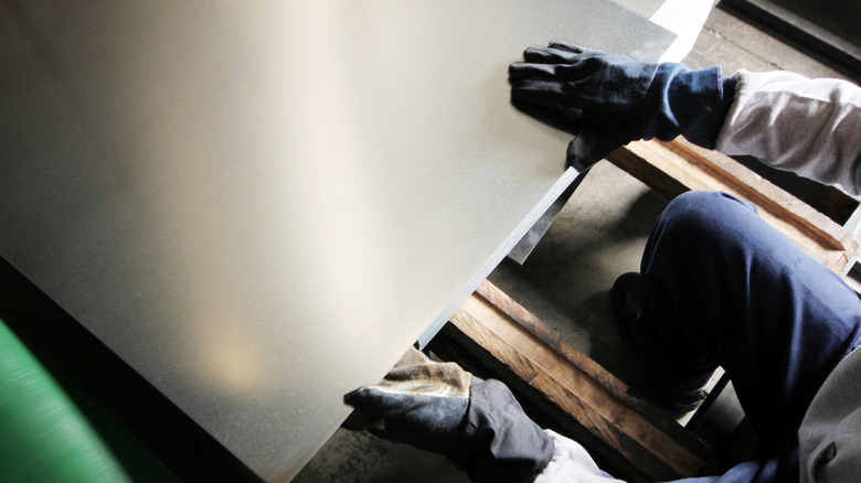 A piece of aluminum sheet metal is being appraised by a worker wearing sturdy gloves.