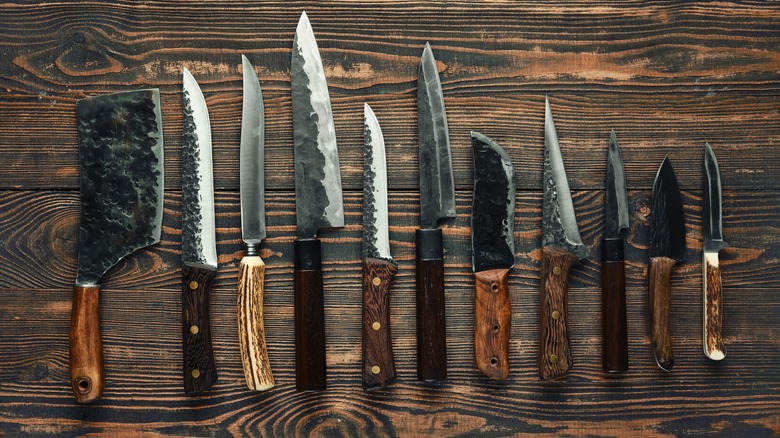 Various kitchen knives are arranged neatly on a rustic wooden table, showcasing different shapes and sizes, including cleaver, utility knives, and chef knives.