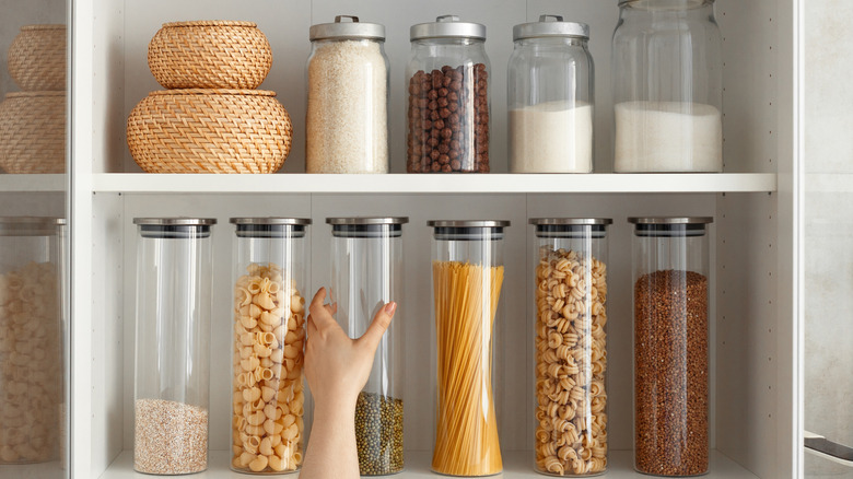 A hand puts back a clear container into a pantry containing a variety of jars with dry ingredients inside