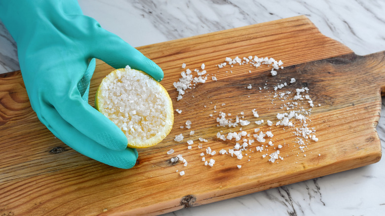 A gloved hand holds half of a lemon that has been rubbed in salt on a wooden cutting board
