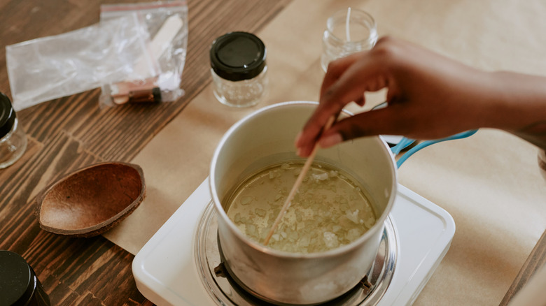 A hand stirring melted wax in a pot on a hot plate near jars prepped for candle making