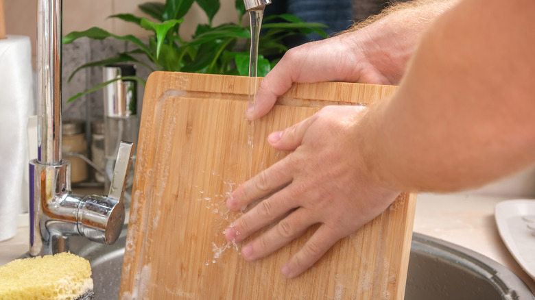 A person's hands holding a wooden cutting board under a running kitchen sink
