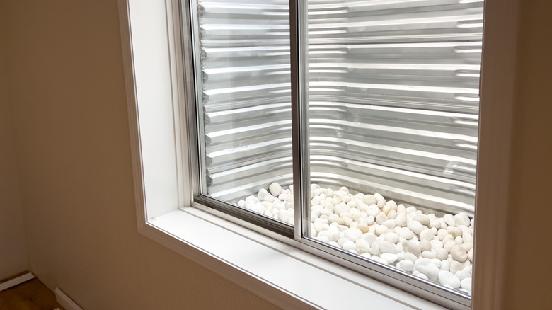 A clear view from a basement window showing a corrugated metal window well lined with smooth white decorative stones at the base.