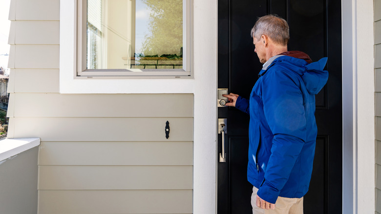 An older man is seen, from behind, entering a code into a smart lock on the door of a home.
