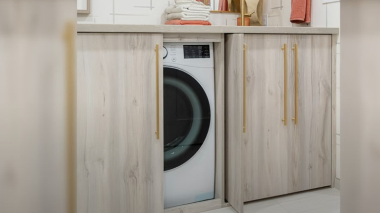 Light wooden cabinetry in a bathroom is pulled aside to reveal a laundry machine inside.