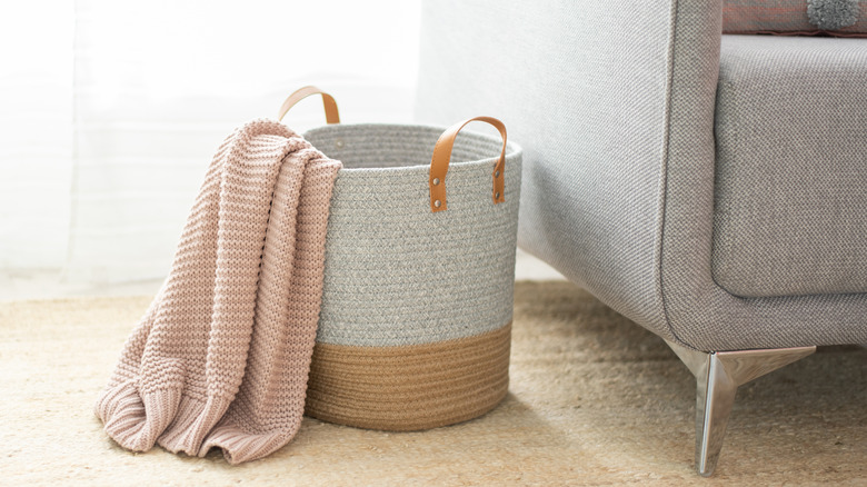 A woven storage basket with a blanket hanging out of it beside a gray sofa.