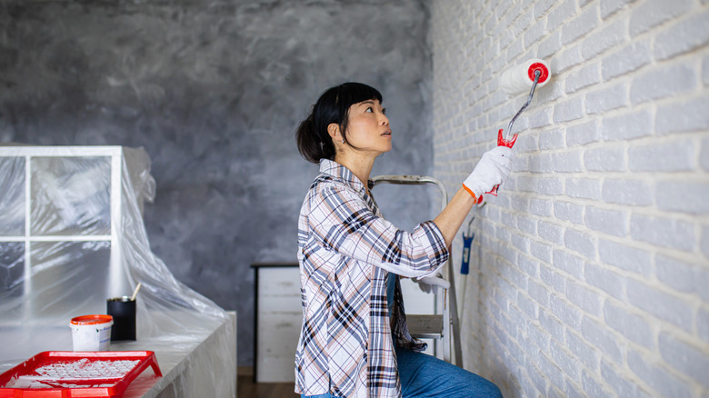 Woman covering her brick with white paint using a roller, with plastic-covered furniture in the background