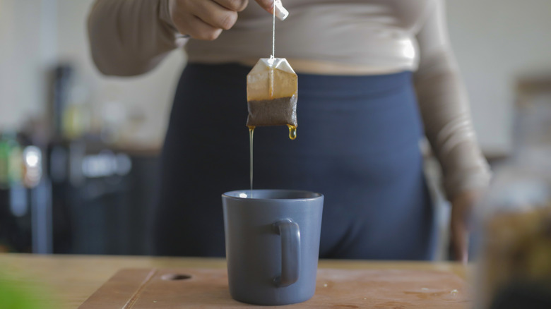 A woman standing behind a blue mug pulling a dripping tea bag out of the mug with one hand