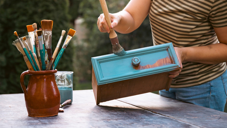 A woman painting a wooden drawer light blue with a thick paint brush outdoors