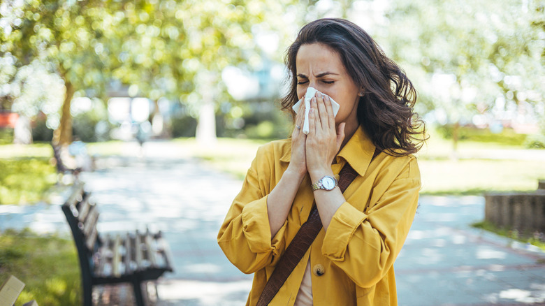 A young caucasian woman sneezes into a tissue while walking through a park in springtime.