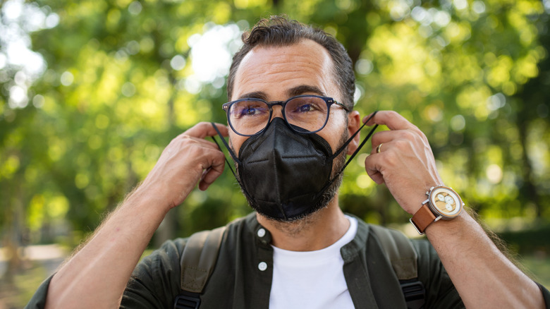 A man wearing glasses and a backpack stands outside and puts on a protective black mask.