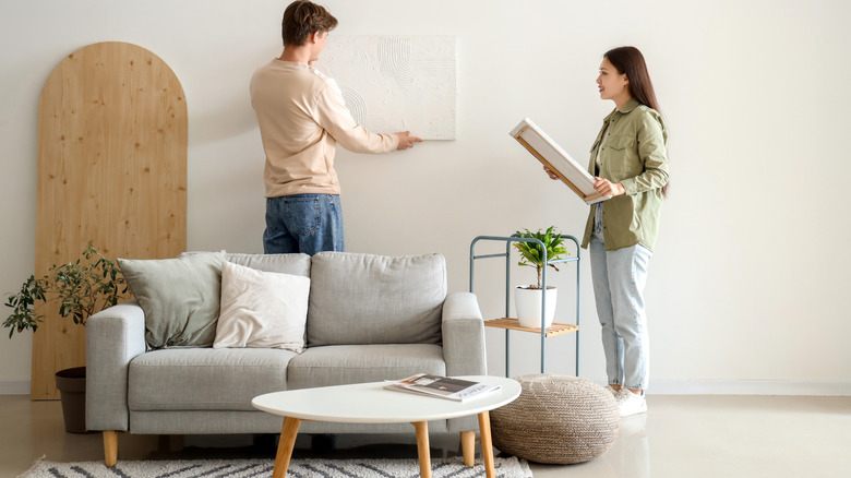 Two people hanging art on the wall behind a sofa