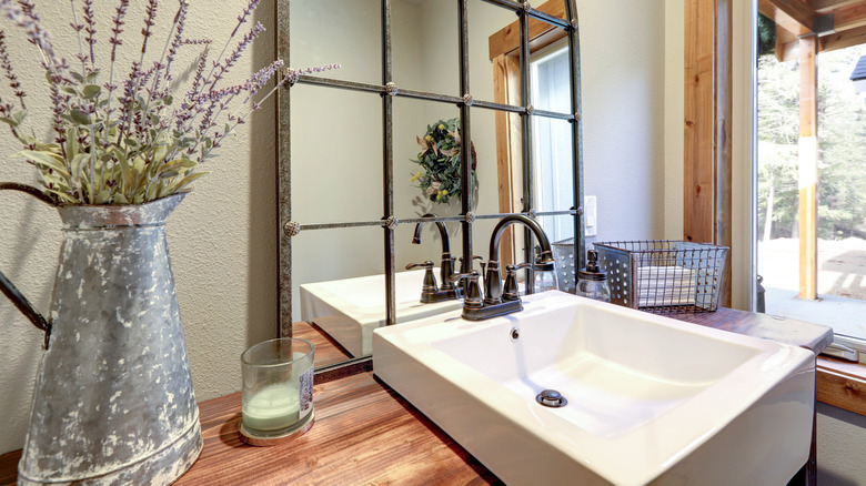 A bathroom vanity and sink with a large mirror and dried flowers and a window in the background.