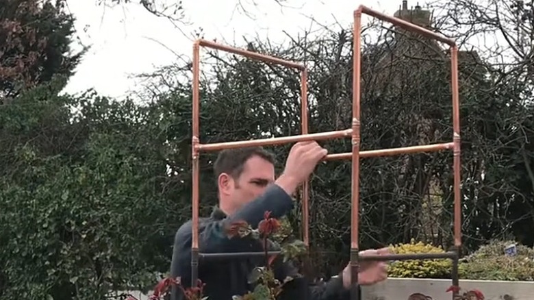 Person building a copper pipe trellis to support roses in the garden.