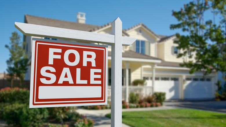 Home for sale with red and white FOR SALE sign in the yard, blurred focus of a yellow home