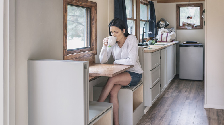Woman sits drinking from a mug at a wooden booth in a tiny home