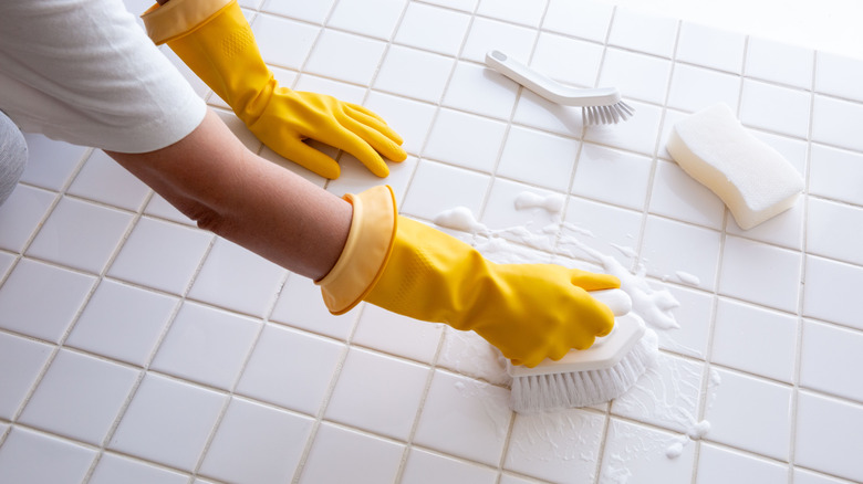A person in yellow gloves is scrubbing white bathroom floor tiles