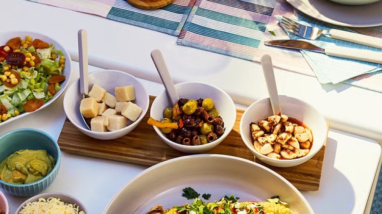 A wooden tray with three white bowls holding olives, cheese, and garlic