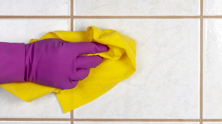 A person wearing gloves wiping white tiles with warm beige grout