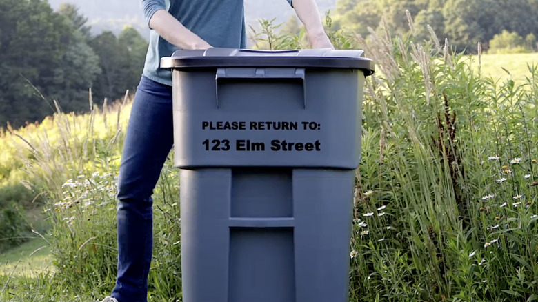 Hands wearing orange gloves take a large black plastic bag full of items out of a green trash can in front of an orange-painted wall.