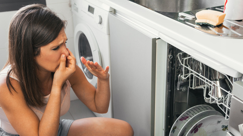 A woman holds her nose in front of an open dishwasher in the kitchen.