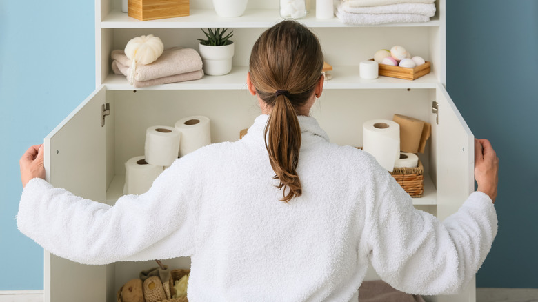 A woman stands in front of an open bathroom cabinet full of toilet paper rolls and other bathroom accessories.