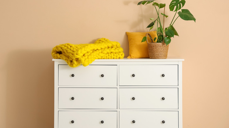 A white dresser with a blanket, pillow and plant on top.