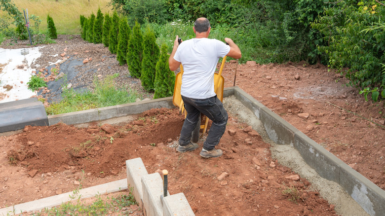 A person dumps soil from a wheelbarrow into the area between two retaining walls in their backyard that's still under construction.