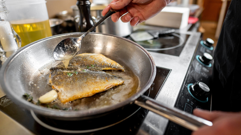 Person using spoon to pour liquid over fish in frying pan