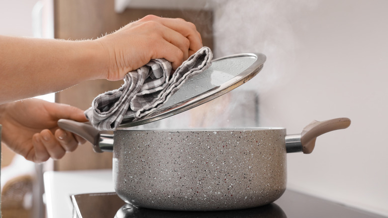 Person using a dish towel to pull the lid off of a hot pot on the oven