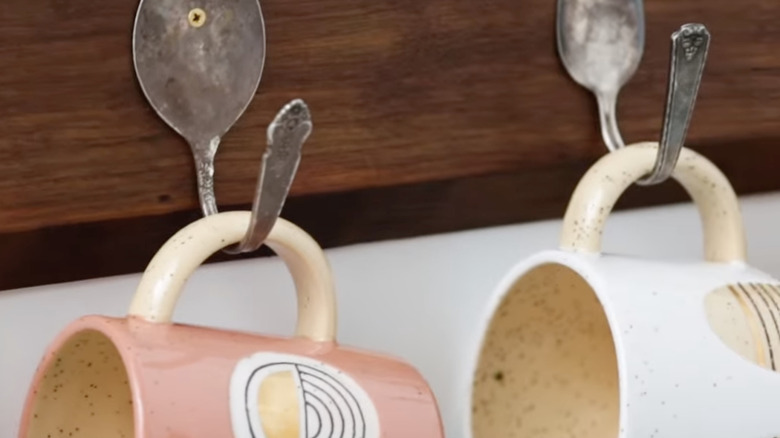 Mugs hang from hooks made from bent spoons attached to a wall-mounted wooden board.